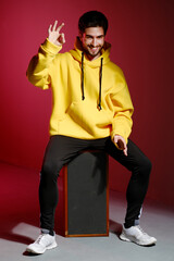 Wall Mural - A young man of 25-30 years old in a yellow sweatshirt sitting on an old music column on pink wall background and showing different emotion.