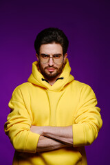 Wall Mural - A young man of 25-30 years in glasses and a yellow sweatshirt emotionally poses on a purple background. 