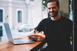© BullRun - Portrait of smiling male writing notes on pages while sitting at wooden table in cozy urban cafe. Bearded young freelancer looking happy and poses for camera using notebook and computer in coffee shop