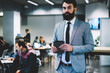 © BullRun - Portrait of confident bearded executive manager dressed in formal wear holding modern touch pad in hands looking at camera while standing in office building with professional it developers