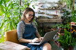 © DimaBerlin - Woman gardener in glasses wear blue jeans overalls, sitting on chair in greenhouse, using laptop after work, communicates on internet with customer surrounded by plants. Home gardening, freelance.