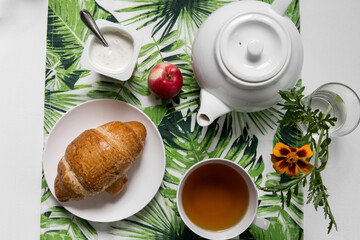 Classic breakfast in a white bowl on a trendy background with a herbal pattern. Croissant, herbal tea, green, flower, fruit, yogurt, apples, healthy, vegetarian.