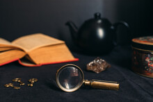 Collection Of Mysterious Items: Old Book, Magnifying Glass, Little Keys, Black Tea Pot, Stone And Tin Box. Black Background.