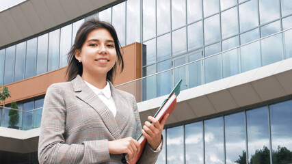 Beautiful smiling confident successful young business woman pretty face looking at camera posing alone near building office, happy millennial girl student with documents professional 