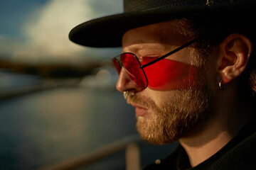 Wall Mural - Side view of trendy fashionable young bearded male wearing stylish hat, pink round sunglasses, earrings and nose piercing enjoying warm sunny weather while walking alone on city embankment