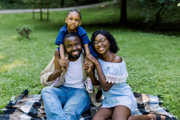 Adorable young African family with cute little daughter, having their weekend time in a city park. Image of cheerful family looking at the camera while picnicking in the park