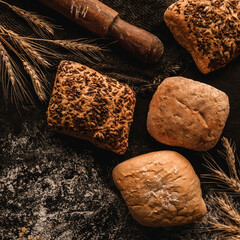 Wall Mural - Different loaf of breads and buns with sesame on dark rustic background with sackcloth, spikelets, rolling pin. Pastries and bread in a bakery, top view