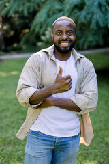 Outdoor casual lifestyle portrait of young African dark skinned man, wearing white t-shirt, beige shirt and jeans, smiling to camera in park nature, showing his thumb up