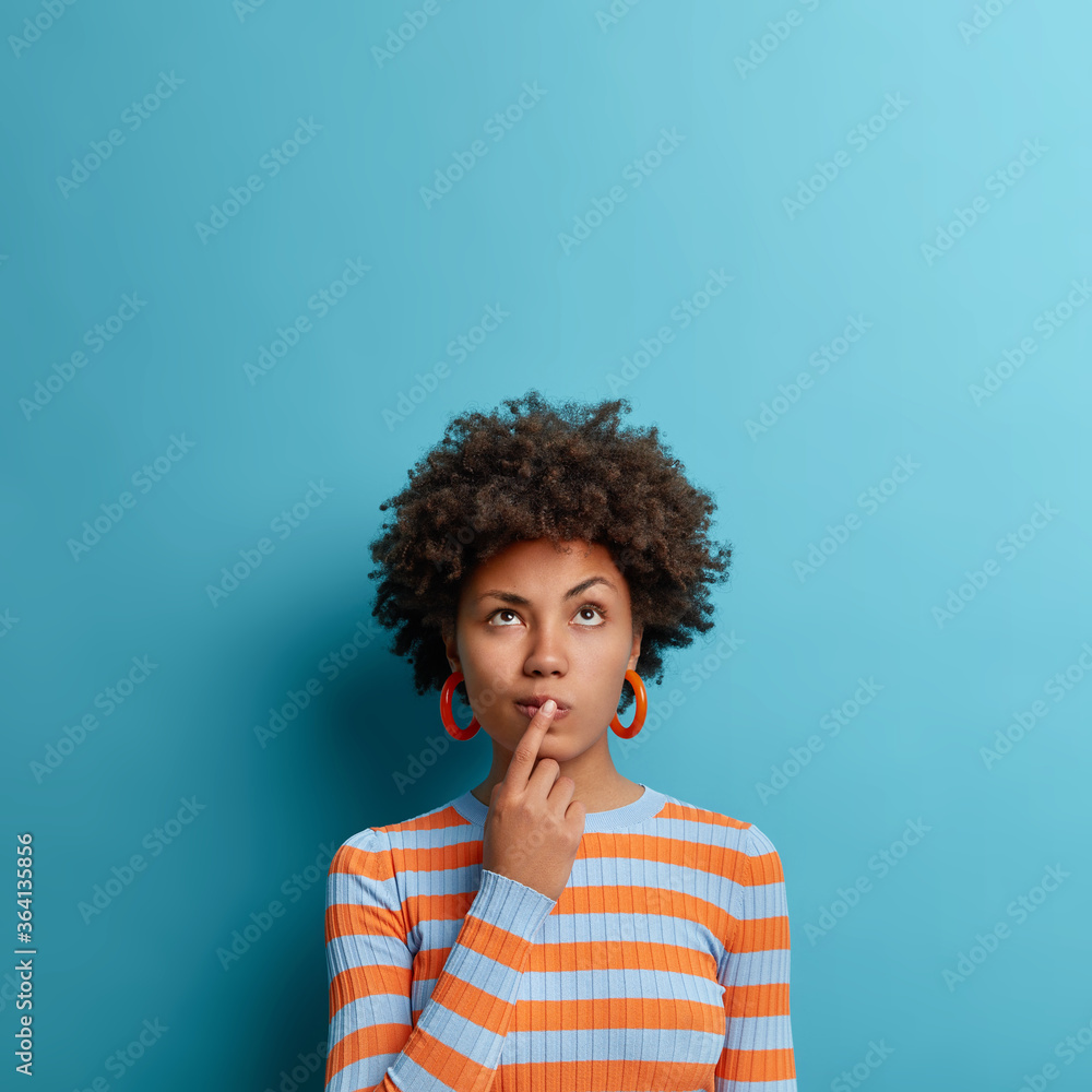 Vertical shot of thoughtful dark skinned woman has idea in mind, keeps finger on lips, looks pensively above, tries to decide how to act, poses against blue background, blank copy space above - obrazy, fototapety, plakaty 