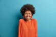 © wayhome.studio  - Upbeat cheerful African American woman laughs with joy, watches funny movie with friend, has carefree expression, talks casually in pub, dressed in orange jumper, isolated on blue background