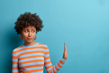 Get Away From Me. Displeased Curly Woman Pulls Hand Aside In Stop Or No Gesture, Being Bothered By Someone, Denies Problems, Ignores Something, Dressed In Casual Striped Jumper, Isolated On Blue Wall
