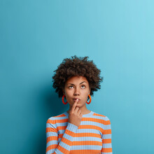 Vertical Shot Of Thoughtful Dark Skinned Woman Has Idea In Mind, Keeps Finger On Lips, Looks Pensively Above, Tries To Decide How To Act, Poses Against Blue Background, Blank Copy Space Above