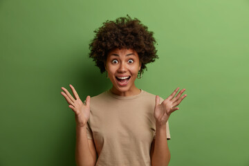 Wall Mural - Joyful very excited cheerful African American woman happy to receive awesome present, raises hands and gestures actively, cannot believe her huge success, dressed in casual wear isolated on green wall