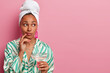 Thoughful lady enjoys drinking martini cocktail after taking shower, looks aside, has well cared complexion, healthy skin, wears dressing gown and towel, isolated on pink studio wall, blank space