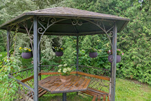 Metal Hexagonal Gazebo With Ornate Elements, With Wooden Benches Around  Perimeter, Wooden Table In Middle, With Vase Of White Flowers On It And Gray Wicker Baskets With Flowers Around, Among Trees