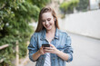 Beautiful blonde teenage girl in denim jacket looks at smartphone smiling. Young woman smiles looking at her smartphone walking down the street