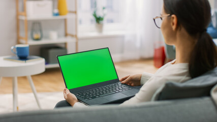 Young Woman at Home Sitting on a Couch Works on a Laptop Computer with Green Mock-up Screen. Girl Using Computer, Browsing through Internet, Watching Content, Chatting in Social Networks with Friends.