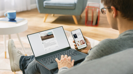 Young Man at Home is Using Laptop Computer, Srufing the Net, Watching News, while Holding Smartphone with Social Media Interface. He's Sitting On a Couch in His Cozy Living Room. Over Shoulder Shot