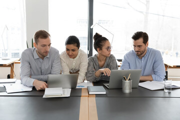 Wall Mural - Concentrated multiracial colleagues sit at desk in office work in groups discuss project using laptops, serious diverse multiethnic employees brainstorm cooperate on computers, teamwork concept