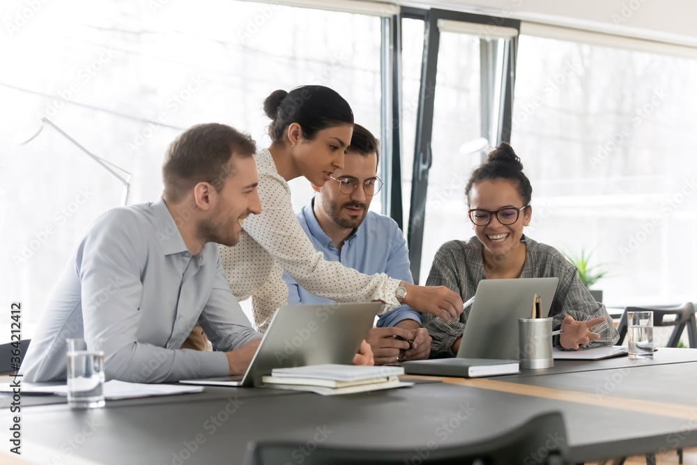 Smiling diverse young colleagues gather at desk in office look at ...