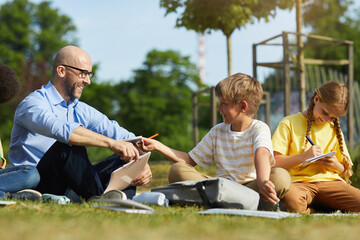 Wall Mural - full length portrait of smiling male teacher talking to teenage boy while sitting on green grass and