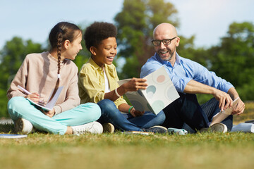 Wall Mural - full length portrait of smiling male teacher talking to children while sitting on green grass and en
