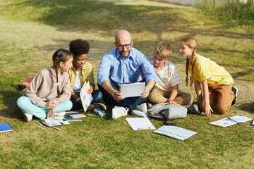 Wall Mural - full length portrait of smiling male teacher talking to group of children while sitting on green gra
