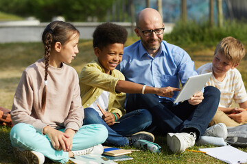 Wall Mural - full length portrait of male teacher talking to group of children while sitting on green grass and e