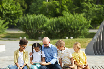 Wall Mural - Front view at multi-ethnic group of children using tablets and smartphones while sitting in row outdoors in sunlight with mature man helping them, copy space