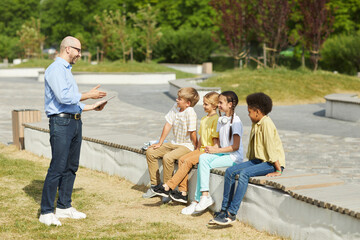Wall Mural - full length portrait of smiling male teacher talking to group of children while enjoying outdoor les