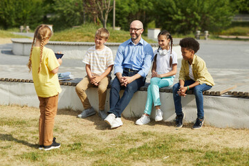 Wall Mural - Full length portrait of smiling male teacher listening to little girl giving presentation while sitting with group of children and enjoying outdoor lesson in sunlight, copy space