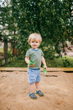 Fototapeta Młodzieżowe - Beautiful little boy in light T-shirt and shorts plays in a sandbox with plastic toys