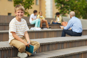 Wall Mural - full length portrait of blonde teenage boy smiling at camera while sitting on bench outdoors with te