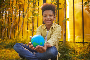 Wall Mural - portrait of smiling african-american boy holding planet in hands while sitting on green grass outdoo