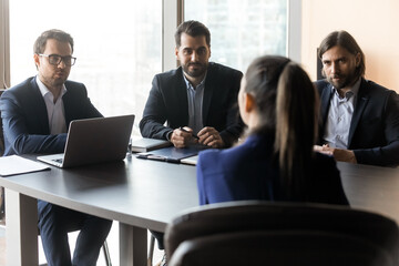 Wall Mural - Group of focused hr managers in formal wear holding job interview with female candidate, sitting together at table. Back view young woman sharing working experience with employers at meeting.