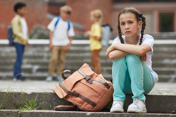 Wall Mural - Full length portrait of sad schoolgirl looking at camera while sitting on stairs outdoors with group of children in background, copy space