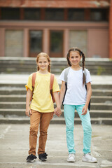 Wall Mural - Vertical full length portrait of two cute schoolgirls with backpacks looking at camera while posing outdoors against school building