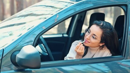 Wall Mural - Young woman sit in car during travelling break. Girl looking into side view mirror and applying lipstick. Make up and cosmetics routine. Take care about her face.