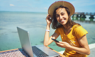 Wall Mural - portrait happy freelancer woman in hat smiling to camera, holding smartphone, working on laptop computer by seashore. Freedom, remote job, freelance, travel and vacation concepts