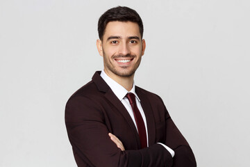 Smiling young manager wearing burgundy suit, white shirt and red tie looking at camera with crossed arms, isolated on gray background