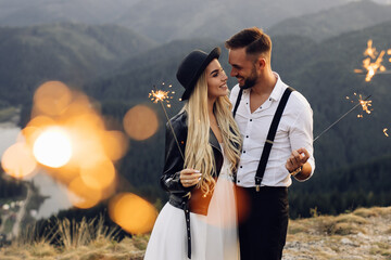 Wall Mural - Beautiful bride and groom with sparklers on the top of the mountain. Wedding photoshoot. Smiling groom and bride. Blonde bride in black jacket .