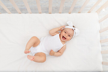 baby girl 6 months old lies in a crib in the nursery with white clothes on her back and laughs, look