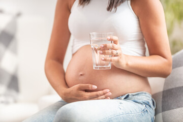 Wall Mural - Pregnant female holding her belly with one hand holds a glass of water in the other hand