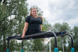 © Михаил Решетников - A beautiful smiling overweight young woman stretches for split on uneven bars outdoors. Fat girl gymnast doing fitness on the sports field. Sport outside on a warm summer day.