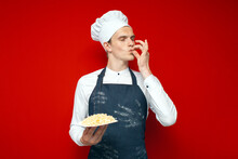 Chef Holds A Dish On A Red Isolated Background And Shows A Sign Of Good Taste, A Kitchen Worker In Uniform Cooked Pasta