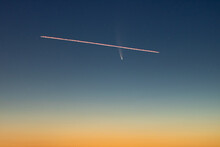 A Plane Flying In Front Of C/2020 F3, Or Comet Neowise, In The Early Morning Sky With A Gradient From Orange Sun Glow To Blue Night Sky. 