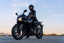 Attractive Girl With Long Hair In Black Leather Jacket And Pants On Outdoors Parking With Stylish Sports Motorcycle At Sunset.