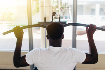 Wall Mural - Strong African American man in sportswear pulling cable with bar while sitting on exercise machine during training in gym.