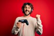 © Krakenimages.com - Young gamer man with curly hair and beard playing video game using joystick and headphones screaming proud and celebrating victory and success very excited, cheering emotion