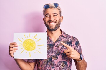 Handsome blond tourist man on vacation wearing sunglasses holding paper with sun draw smiling happy pointing with hand and finger